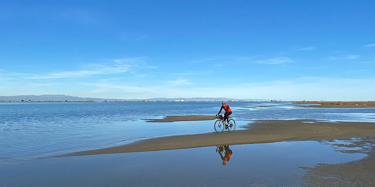 Ciclista con chaleco rojo y bolsas de bikepacking pedaleando junto a la orilla del mar en la Barra del Trabucador